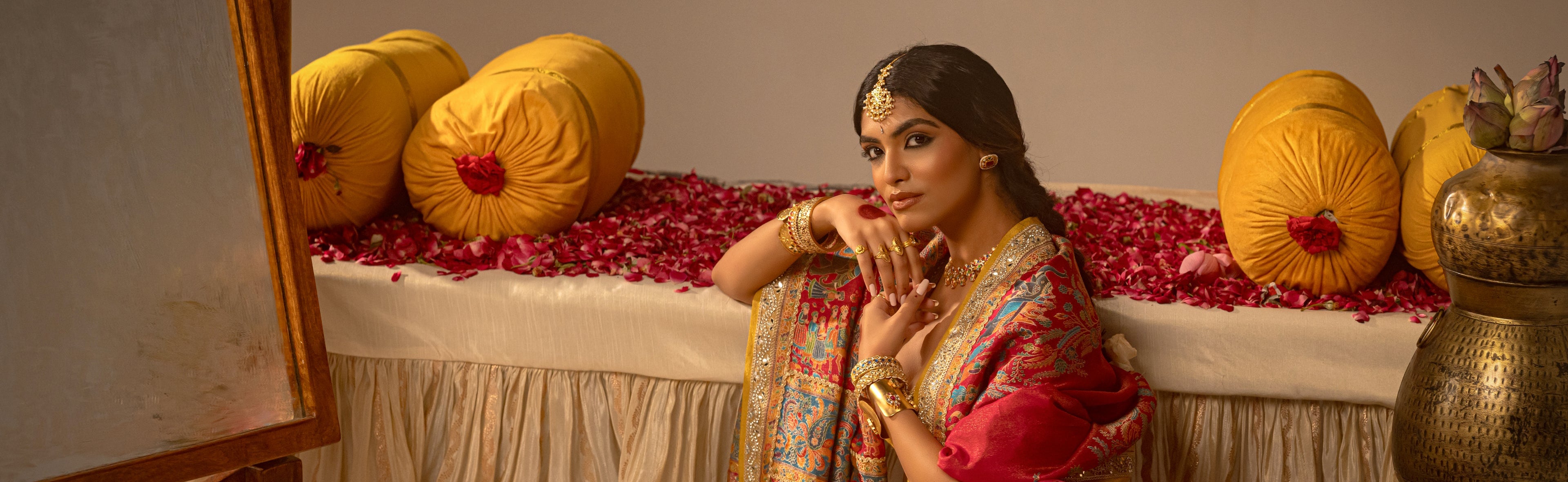 Woman in traditional attire sitting on a decorated bed with yellow cushions and red flowers.